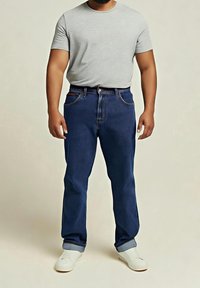 Homme portant une chemise à manches courtes gris clair, un jean bleu foncé avec des revers et des chaussures blanches sans lacets, debout sur un fond clair uni.