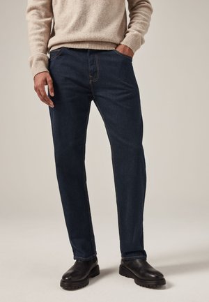 Hombre que lleva vaqueros azul oscuro de pernera recta, suéter beige y botas de cuero negras, de pie con una mano en el bolsillo.