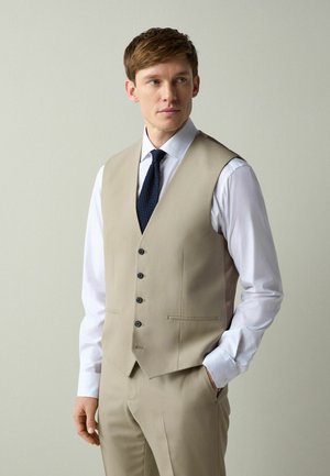 Man wearing beige suit vest and pants with white shirt and dark blue tie, standing against plain light background.