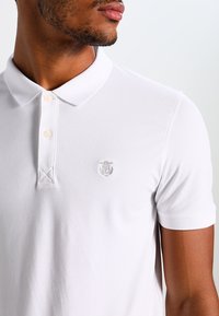 Bílá polokošile s tříknoflíkovou légou, krátkými rukávy a decentním vyšívaným logem na hrudi. Texturovaný povrch látky.