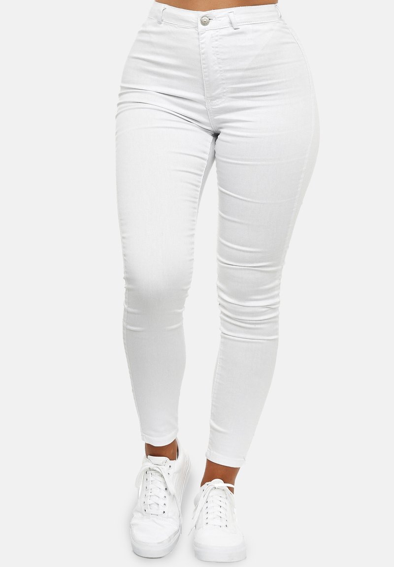 Mujer vestida con jeans blancos de tiro alto y corte ajustado, y zapatillas blancas, de pie contra un fondo liso claro.