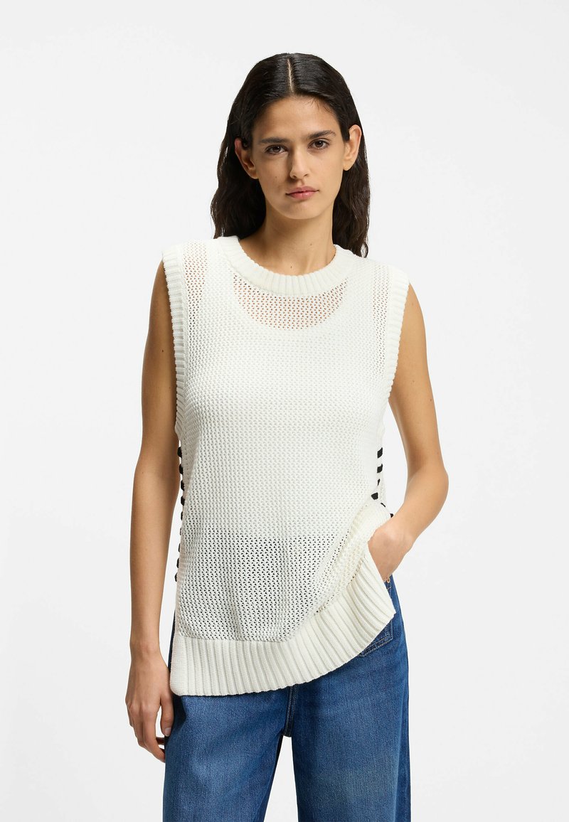 Femme aux longs cheveux foncés portant un haut en maille blanc sans manches et un jean bleu, debout avec une main dans la poche devant un fond uni.