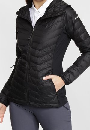 Vrouw draagt een zwarte doorgestikte jas met ritssluiting, capuchon en zijzakken, gecombineerd met grijze broek en een wit overhemd met kraag eronder.