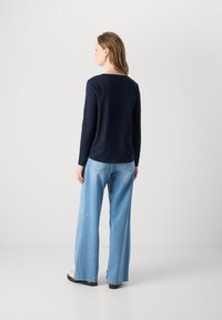 Marineblaue Langarmbluse mit Rundhalsausschnitt, kombiniert mit hellblauen weiten Jeans und klobigen schwarzen Schuhen, von hinten betrachtet.