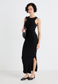 Robe maxi noire à côtes avec une silhouette ajustée, design sans manches, fente sur le côté, associée à des ballerines noires à bride arrière et à un petit sac.