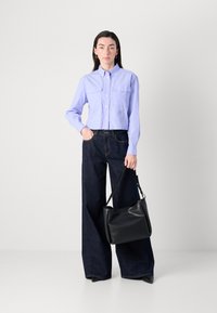 Camicia button-up viola chiaro con tasche a patta, abbinata a jeans scuri a gamba larga. Una borsa nera a forma curva è tenuta in una mano.