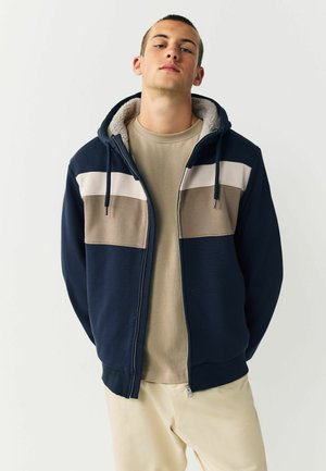 Marineblauer Reißverschluss-Hoodie mit hellgrauen und braunen horizontalen Streifen, fleecegefütterter Kapuze und Kordeln. Getragen über einem beigen Hemd.