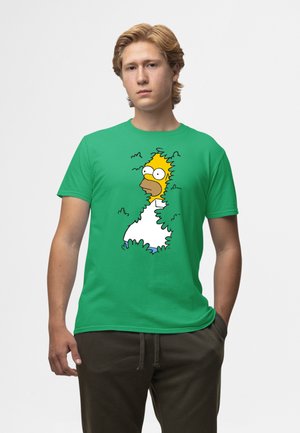 Zelené bavlněné tričko s karikaturou částečně skrytou za listím, s živými barvami a uvolněným střihem.