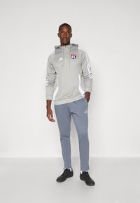 Grå Adidas hoodie med hvide accenter og logo, parret med lysegrå joggerbukser og sorte træningssko på en ensfarvet baggrund.