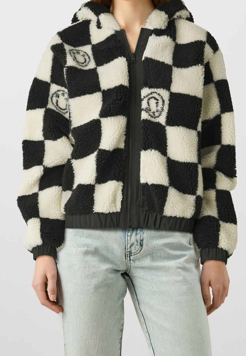 Persona que lleva una chaqueta de forro polar a cuadros en blanco y negro con cremallera y jeans de mezclilla de color azul claro, de pie contra un fondo liso.