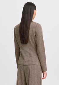 Blazer marrone a spina di pesce con maniche lunghe e silhouette aderente, caratterizzato da una texture liscia e un design posteriore sartoriale.