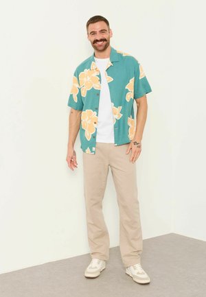 Mann mit Schnurrbart lächelt, trägt ein türkisfarbenes Kurzarmhemd mit gelben Blumen über einem weißen T-Shirt, beige Hose und weiße Sneakers, steht drinnen.