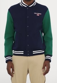 Marinblå och grön varsityjacka med vita ränder på kragen och ärmslut, med framknäppning och sidofickor.