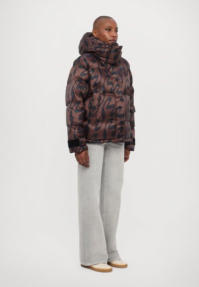 Brown patterned puffer jacket with high collar, elastic cuffs, and front buttons paired with light grey flared jeans and beige shoes.