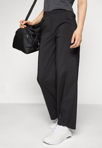 Sorte, lette, elastiske bukser med løs passform, kombinert med en grå topp og hvite joggesko. Svart treningsbag holdt i den ene hånden.