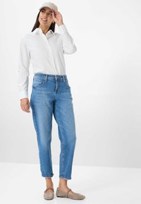 Hellblaue Jeans im entspannten Schnitt, ein weißes Hemd mit Knöpfen und eine beige Baseballkappe. Graue flache Schuhe runden das Outfit ab.