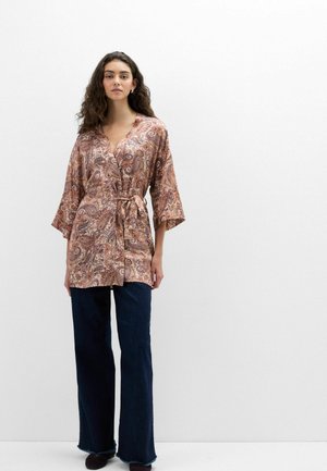 Frau mit langen lockigen Haaren, die eine rosa Tunika mit Paisley-Muster und Taillenbindegürtel sowie dunkle, weit geschnittene Jeans trägt, steht vor einem schlichten weißen Hintergrund.