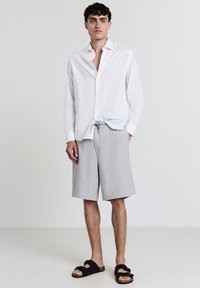 Jeune homme portant une chemise blanche à boutons, un short gris clair à cordon et des sandales noires à enfiler, se tenant devant un fond uni.