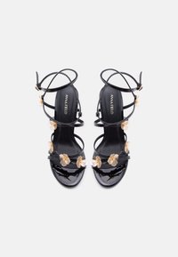 Μαύρα σανδάλια από δερματίνη με πολλαπλούς ιμάντες, διακοσμημένα με χρυσές ανθισμένες λεπτομέρειες και διαφανή ιμάντα. Η σόλα είναι κομψή και ανοιχτή μπροστά.