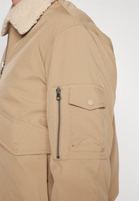 Chaqueta beige con un cuello de borrego texturizado. Presenta un bolsillo con cremallera en la parte superior del brazo y un bolsillo de botón a presión debajo de este.