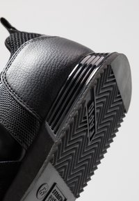 Semelle de chaussure noire avec motif de bande de roulement en zigzag, empeigne en cuir noir texturé, et détail de talon empilé brillant sur un fond uni.