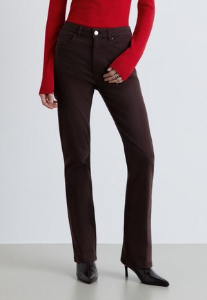 Donna che indossa pantaloni dritti marrone scuro, stivali neri a punta con tacco alto e una maglia rossa aderente a maniche lunghe, in piedi davanti a uno sfondo semplice.