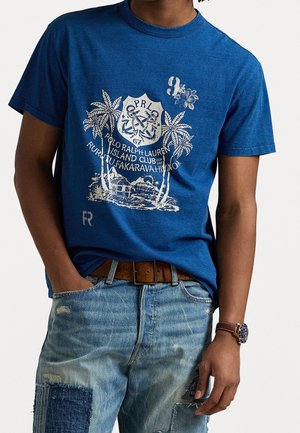 Mężczyzna w niebieskiej koszulce graficznej z nadrukiem palmy i klubu wyspiarskiego, w połączeniu z jasnymi dżinsami z łatami oraz brązowym skórzanym paskiem i zegarkiem.