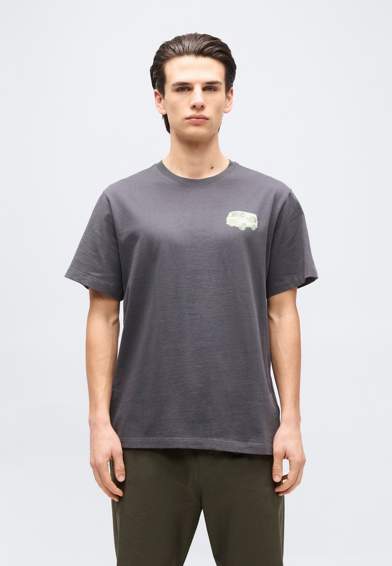 Junger Mann mit dunklem Haar trägt ein dunkelgraues T-Shirt mit einer kleinen hellfarbenen Van-Grafik auf der linken Brust vor einem einfarbigen Hintergrund.