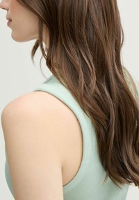 Mujer con cabello largo castaño, vistiendo un top sin mangas de color verde claro con textura acanalada, mostrada de perfil y por detrás contra un fondo sencillo.