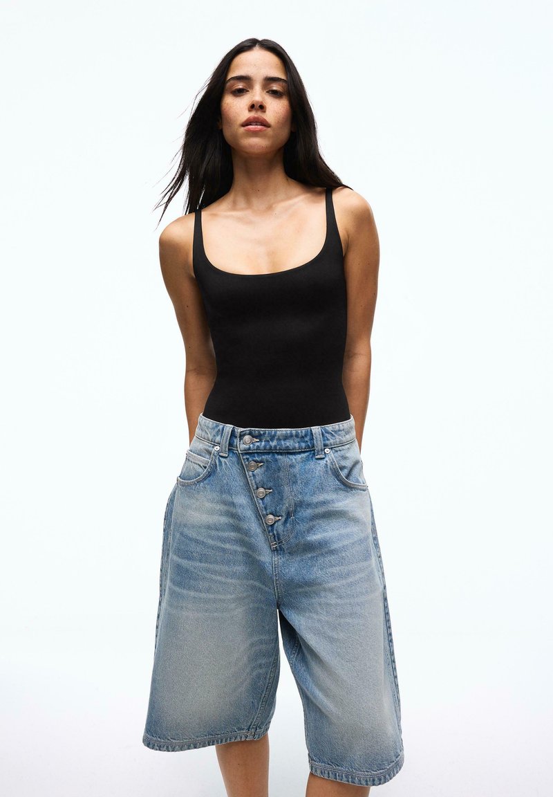 Femme aux longs cheveux foncés portant un haut noir sans manches et un short en jean ample, taille haute, avec des détails de boutons.