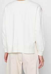 Sudadera blanca de algodón con hombros caídos, puños y dobladillo acanalados. Presenta un ajuste relajado y detalles de costura sutiles. Llevada sobre unos pantalones beige.