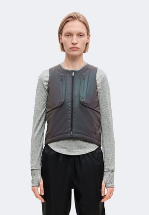 Une femme porte un gilet foncé sans manches, à fermeture éclair sur le devant et muni de poches, par-dessus un haut gris à manches longues et un pantalon noir, sur un fond uni.