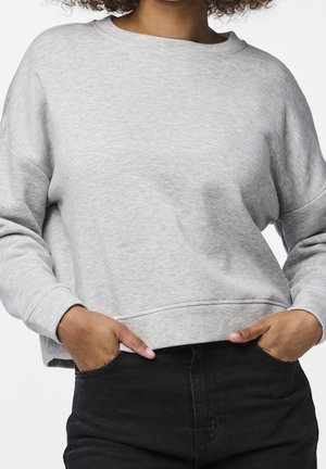 Persoon die een lichtgrijs sweatshirt met lange mouwen draagt, met handen in de zakken van een zwarte spijkerbroek tegen een witte achtergrond.