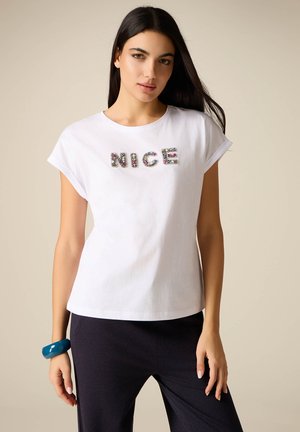 Junge Frau mit langen dunklen Haaren, die ein weißes T-Shirt mit dem dekorativen Schriftzug "NICE" und schwarze Hosen trägt, posiert vor einem schlichten Hintergrund.