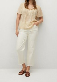 Woman wearing a beige textured blouse, white cropped pants with frayed hems, and brown flat sandals, standing against a plain background.