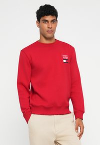 Sweat-shirt rouge avec un col rond, des poignets et une base côtelés. Logo brodé sur le côté gauche de la poitrine avec des accents bleu marine et blanc.