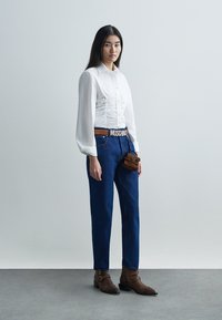 Biała koszula z długim rękawem z marszczeniami, niebieskie dżinsowe spodnie, brązowy pasek z napisem "Moschino", brązowe botki za kostkę, mała brązowa torebka na ramię.