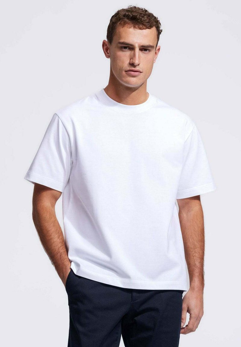 Witte katoenen t-shirt met een ronde hals, korte mouwen en een losvallende pasvorm, gecombineerd met marineblauwe broek. Moeilijke textuur, minimaal ontwerp.