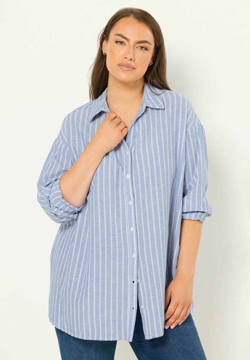 Vrouw met lang bruin haar, gekleed in een losse lichtblauwe blouse met verticale strepen en donkere spijkerbroek, poserend tegen een effen witte achtergrond.