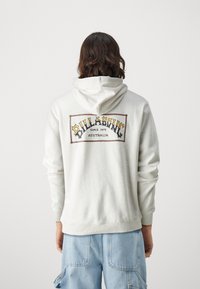 Ανοιχτό γκρι φούτερ με μεγάλο γραφικό τύπωμα στο πίσω μέρος. Μαλακή υφή, με το όνομα της μάρκας "Billabong" και "Since 1973" σε πολύχρωμο κείμενο.