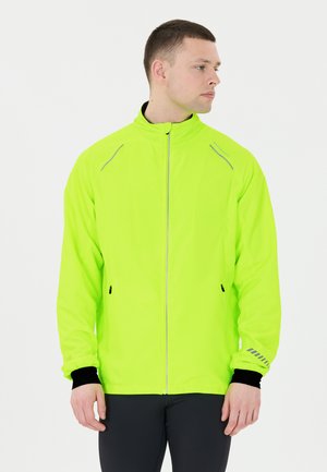 Neon grøn letvægtsjakke med lynlås foran, refleksstriber og sorte manchetter. Har to sidelommer og en stående krave.