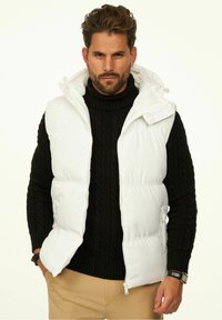 Weiße Steppweste mit Kapuze über einem schwarzen Strickpullover mit Zopfmuster. Verfügt über einen Frontreißverschluss und seitliche Taschen, mit glatter, glänzender Textur.