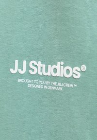 Miętowozielona tkanina o gładkiej fakturze z białym wyhaftowanym napisem: „JJ Studios” oraz mniejszym tekstem poniżej wskazującym pochodzenie marki.