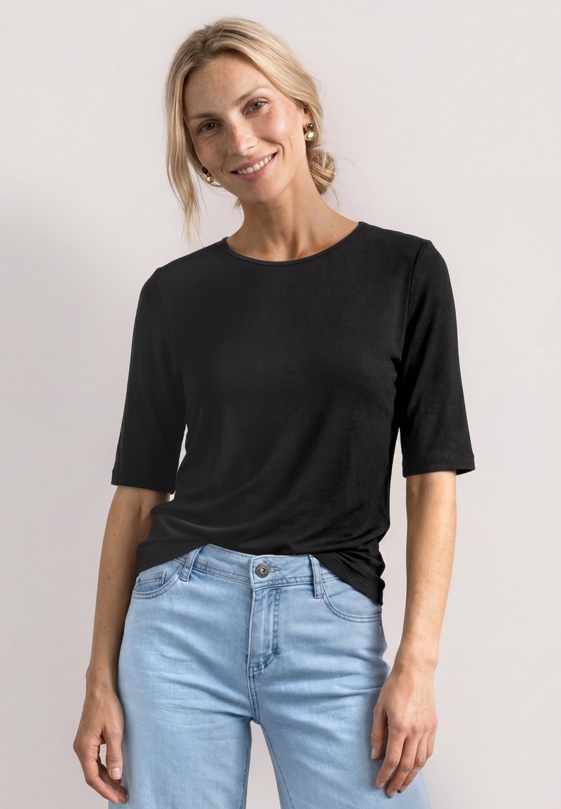 Femme aux cheveux clairs vêtue d’un t-shirt noir à manches courtes et d’un jean bleu clair, souriant sur un fond clair uni.