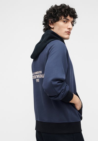 Fiatal férfi göndör hajjal, sötétkék és fekete kapucnis pulóvert visel, amelyen szöveg található a hátoldalon, zsebre tett kézzel, a vállán áttekintve, egyszínű háttér előtt.