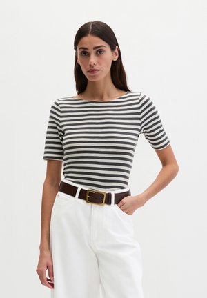 Woman with long dark hair wearing a black and white striped short-sleeve top, white pants, and a dark brown belt with a gold buckle.