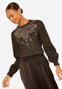 Črna bluza z dolgimi rokavi z teksturiranim zaključkom in zapletenimi dekoracijami iz perlic in sequins na ovratniku, v kombinaciji s črnimi hlačami.
