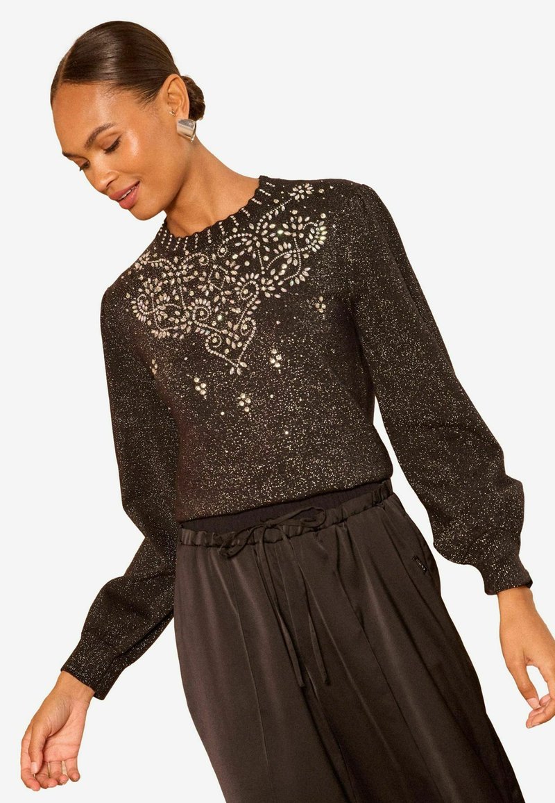 Črna bluza z dolgimi rokavi z teksturiranim zaključkom in zapletenimi dekoracijami iz perlic in sequins na ovratniku, v kombinaciji s črnimi hlačami.
