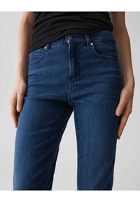 Jean en denim bleu foncé avec une coupe ajustée, style cinq poches, fermeture à bouton unique et texture subtile. Couture visible le long des coutures.
