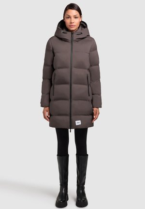 Grijze pufferjas met een capuchon, voorzien van een ritssluiting aan de voorkant, twee zijzakken en elastische accenten. Gecombineerd met zwarte leggings en hoge zwarte laarzen.